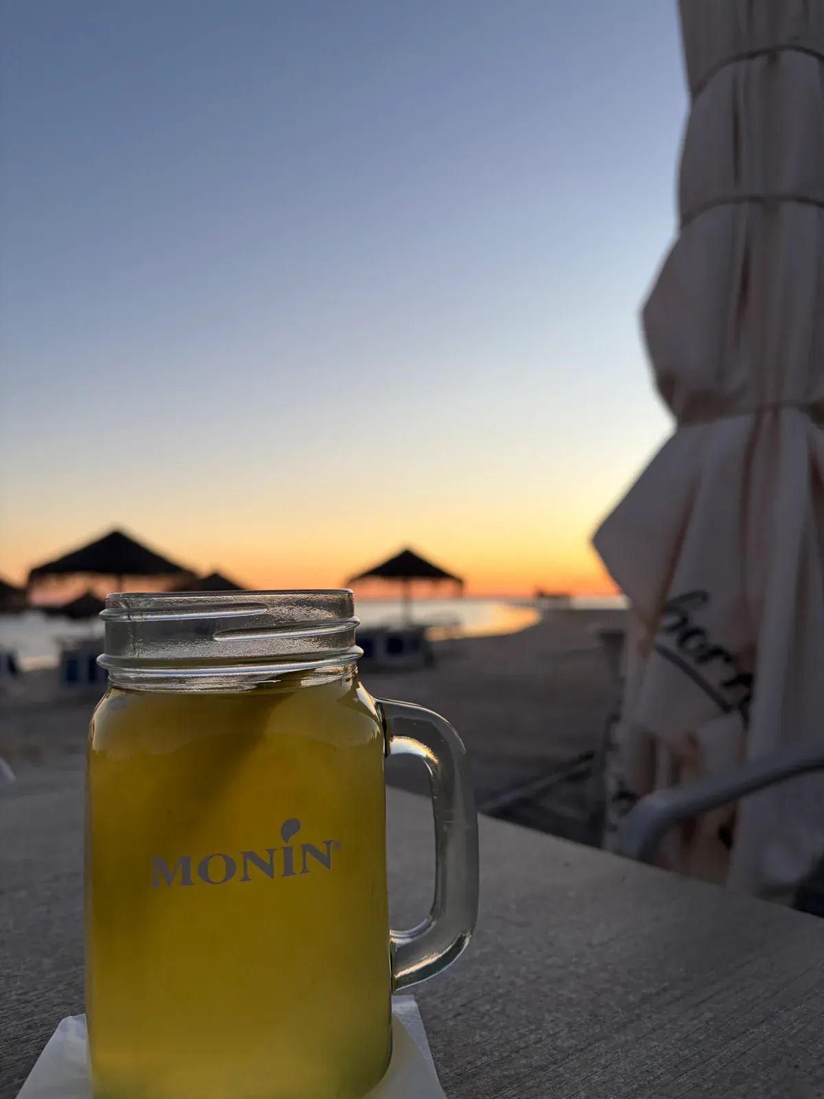 A glass mason-jar mug of a yellow drink on a beach-bar table at dusk, thatched parasols and a furled umbrella silhouetted against an orange sunset over the water