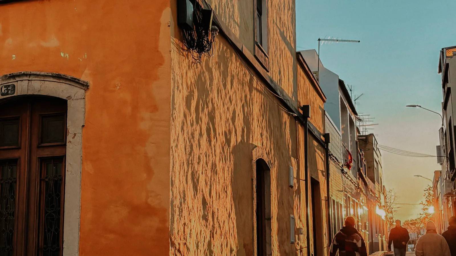 A narrow village street in low backlit sun, ochre and white rendered houses on both sides, several figures in winter coats walking away from the camera