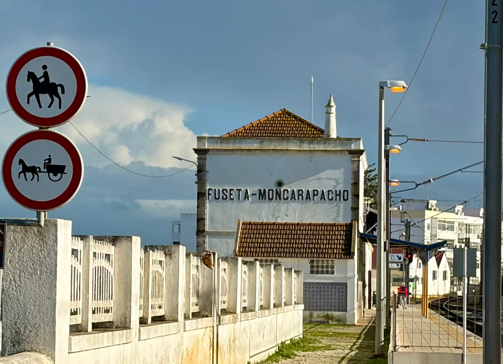 The whitewashed Fuseta-Moncarapacho station building seen along the platform, its name painted across the gable under overhead wires