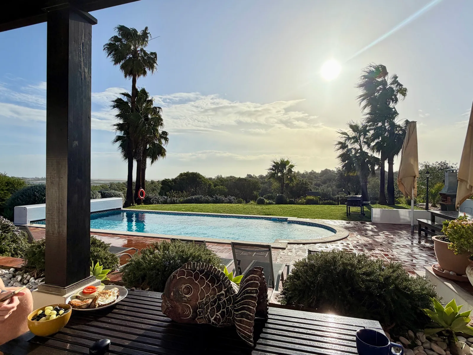A breakfast of eggs and toast beside a bowl of olives on a wooden terrace table, with a swimming pool, palm trees and the lagoon beyond in low morning sun