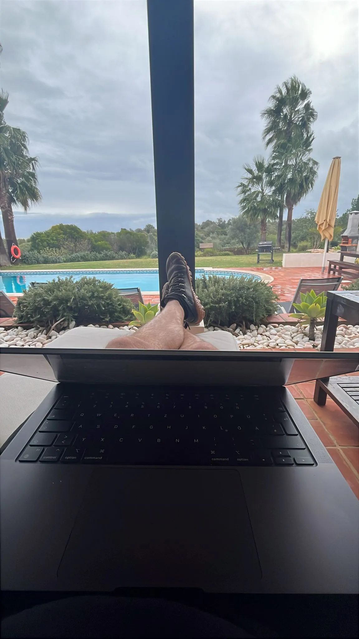 A laptop with a German keyboard layout open on someone's lap, feet up on a cushion, a pool and palm trees under grey cloud beyond