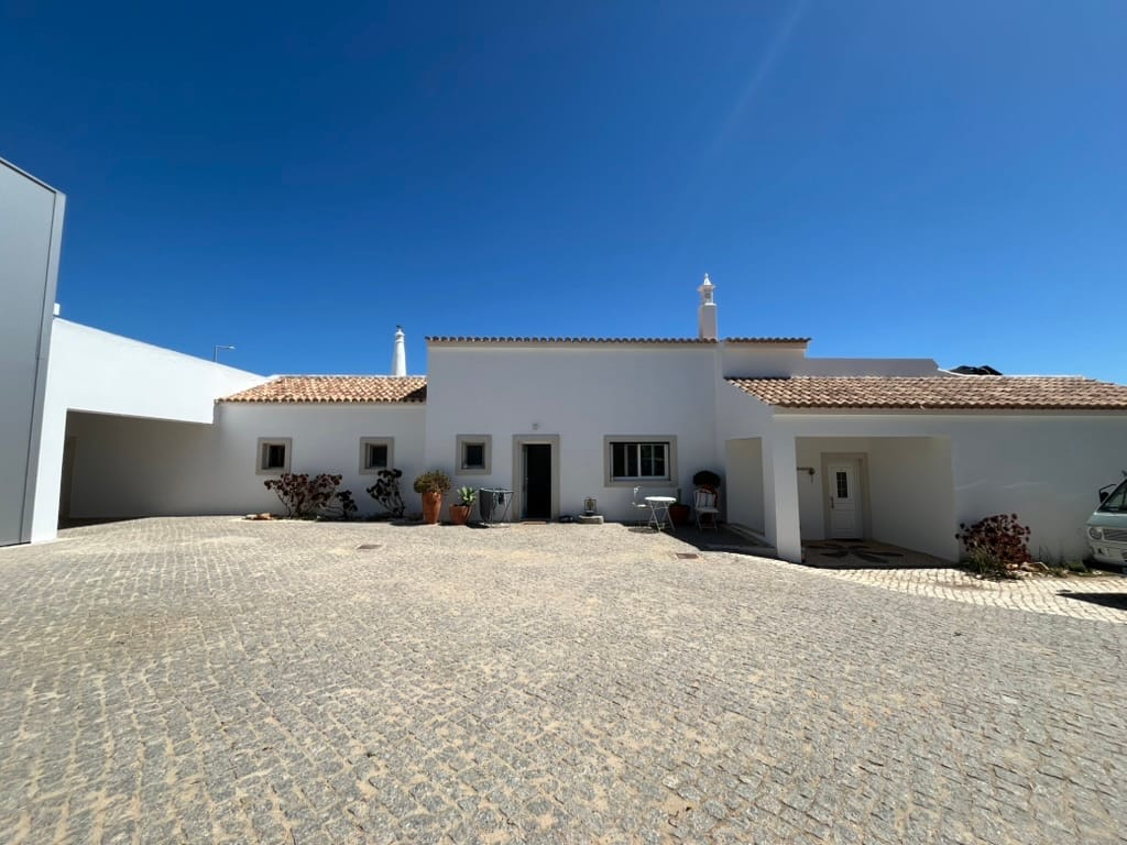 A low whitewashed house with a terracotta roof and traditional Algarve chimneys, seen across a cobbled forecourt under a deep blue sky